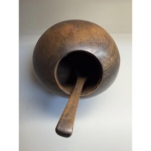 May include: A dark brown, spherical wooden bowl with a smooth, rounded exterior and a wide, circular opening at the top. A flat, rectangular wooden handle extends outward from the interior base of the bowl, protruding through the opening.