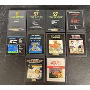 May include: A collection of ten rectangular Atari 2600 video game cartridges arranged in three rows. The black plastic cartridges feature various labels with game titles including Combat, Space Jockey, Video Pinball, Football, and Super Breakout.