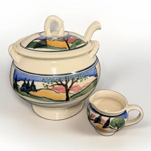 May include: A cream-coloured ceramic soup tureen with a matching lid and ladle, featuring a hand-painted landscape design with trees, hills, and blue sky. A smaller matching cream-coloured ceramic creamer sits beside the tureen.