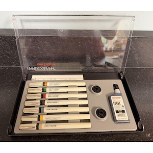 May include: A set of Koh-I-Noor Rapidograph pens in a clear plastic case. The set includes several pens with different colored tips, two ink bottles, and two ink reservoirs. The pens are arranged in a gray tray.