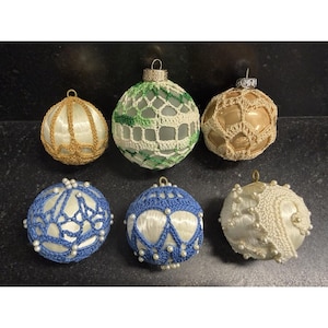 May include: Six spherical Christmas ornaments featuring various designs. The top row includes a gold-wrapped ball, a green and white crocheted ball, and a tan crocheted ball. The bottom row features two blue and white crocheted balls with small white beads and a white ball with white crochet and beads.