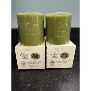 May include: Two green pillar candles, labeled "Pome Green," sit atop their boxes. The boxes are white with the text "Colonial Pillar" and "Colonial Candle Co. of Cape Cod, Inc." printed on them. The candles are cylindrical and wrapped in clear plastic.