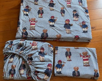 May include: A bedding set featuring a light blue and white pinstriped pattern with a repeating print of teddy bears dressed in various outfits, including sweaters, blazers, and polo shirts, across a flat sheet, a fitted sheet, and a pillowcase.