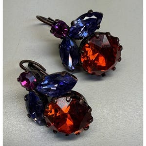 May include: A pair of drop earrings featuring a large, round, faceted orange stone, two marquise-cut blue stones, and a small, round, faceted purple stone. The stones are set in a dark metal frame with a curved hook for attachment to the ear.