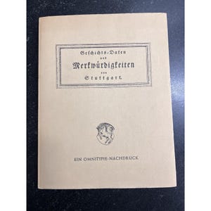 May include: A vintage book with a tan cover featuring German text within a rectangular frame. The text reads "Geschichts-Daten und Merkwürdigkeiten von Stuttgart." Below the text is a small emblem and the text "EIN OMNITYPIE-NACHDRUCK."