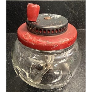 May include: An antique glass jar with a red wooden handle and lid. The jar has a metal top with a crank and a clear glass body with embossed floral designs. The jar's lid is red, and the handle is also red.