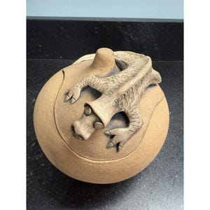 Puede incluir: Un recipiente de cerámica de color beige con tapa, con una detallada escultura de dragón en la parte superior. El dragón tiene escamas texturizadas y está posicionado sobre la tapa redondeada. El recipiente es redondo y tiene una superficie lisa.