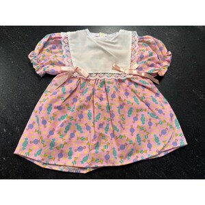 Puede incluir: Un vestido de manga corta con fondo rosa y un patrón repetitivo de caramelos en color verde azulado y morado. El corpiño tiene un panel blanco con ribete de encaje y dos cintas de color rosa claro atadas en la cintura a ambos lados.