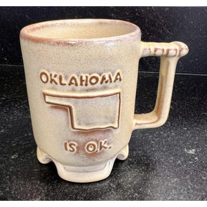 May include: Tan ceramic mug with the word "OKLAHOMA" and a state outline in brown. The phrase "IS OK." is below the state outline. The mug has a unique handle and three small feet.
