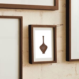 May include: A framed wall art print featuring a centered illustration of a brown, diamond-shaped bottle with a long, slender neck. The artwork is displayed in a dark wood frame, mounted on a textured, off-white brick wall alongside other frames.