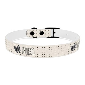 Könnte beinhalten: Ein cremefarbenes Hundehalsband mit schwarzen Punkten und dem Wort "BIKU" in fetter Schrift. Das Halsband hat eine schwarze Schnalle und eine Schleifen-Grafik. Für Haustiere.