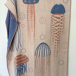 K&ouml;nnte beinhalten: Beige &Uuml;berwurfdecke mit einem sich wiederholenden Muster aus blauen und braunen Quallen und Sanddollars. Die Decke hat einen blauen Rand und Fransen am unteren Rand. Die Quallen haben gepunktete und gestreifte Designs.
