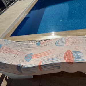 K&ouml;nnte beinhalten: Ein hellrosa Strandtuch mit einem Quallenmuster in Koralle und Blau, drapiert &uuml;ber einer wei&szlig;en Sonnenliege. Das Handtuch hat einen Fransenrand und befindet sich in der N&auml;he eines Swimmingpools.