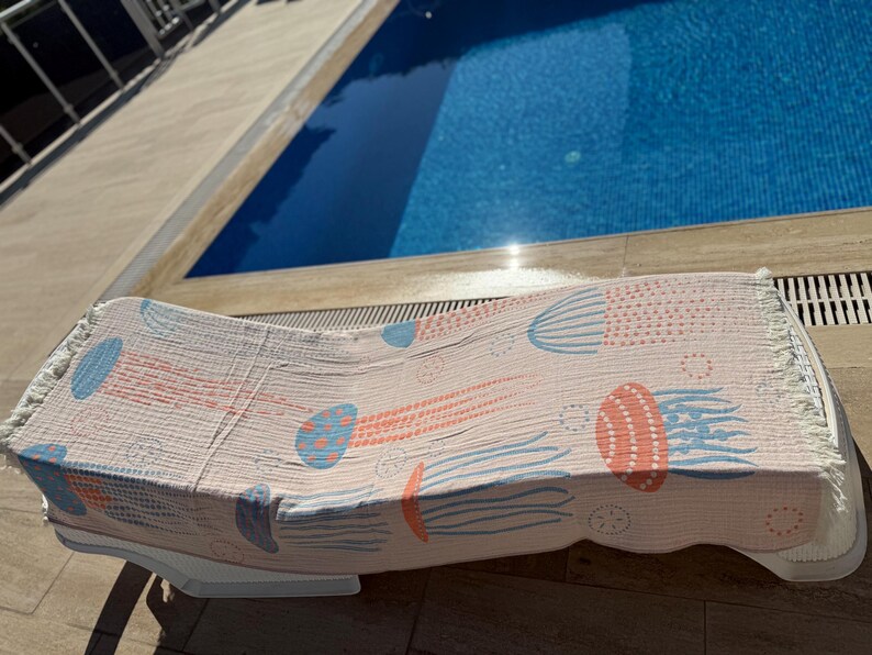 K&ouml;nnte beinhalten: Ein hellrosa Strandtuch mit einem Quallenmuster in Koralle und Blau, drapiert &uuml;ber einer wei&szlig;en Sonnenliege. Das Handtuch hat einen Fransenrand und befindet sich in der N&auml;he eines Swimmingpools.