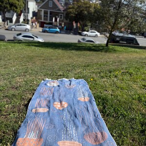 K&ouml;nnte beinhalten: Eine blaue Stranddecke mit einem sich wiederholenden Muster aus korallen- und wei&szlig;en Formen. Die Decke hat einen Fransenrand und ist auf einer grasbewachsenen Fl&auml;che ausgebreitet. Der Hintergrund zeigt ein Wohnviertel mit H&auml;usern und Autos.