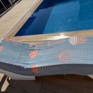 K&ouml;nnte beinhalten: Hellblaues Strandtuch mit Quallenmuster in Koralle und Wei&szlig;. Das Tuch liegt auf einer wei&szlig;en Sonnenliege neben einem Swimmingpool. Mit Fransen.