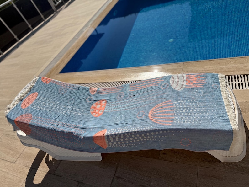 K&ouml;nnte beinhalten: Hellblaues Strandtuch mit Quallenmuster in Koralle und Wei&szlig;. Das Tuch liegt auf einer wei&szlig;en Sonnenliege neben einem Swimmingpool. Mit Fransen.