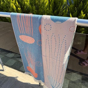 K&ouml;nnte beinhalten: Zwei Strandt&uuml;cher h&auml;ngen an einem Metallgel&auml;nder. Eines ist hellblau mit orangefarbenen und wei&szlig;en Designs, das andere ist rosa mit blauen und wei&szlig;en Designs. Beide haben Fransen und sind im Freien.