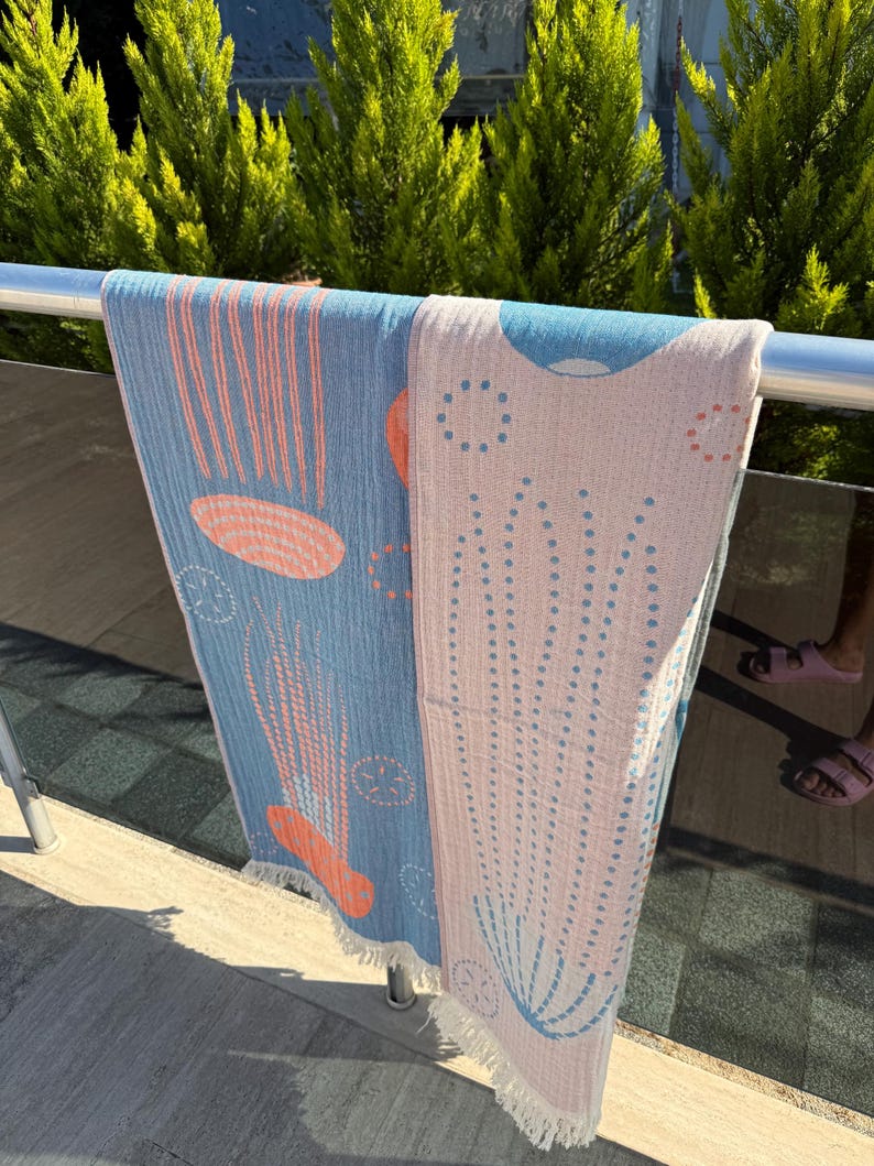 K&ouml;nnte beinhalten: Zwei Strandt&uuml;cher h&auml;ngen an einem Metallgel&auml;nder. Eines ist hellblau mit orangefarbenen und wei&szlig;en Designs, das andere ist rosa mit blauen und wei&szlig;en Designs. Beide haben Fransen und sind im Freien.