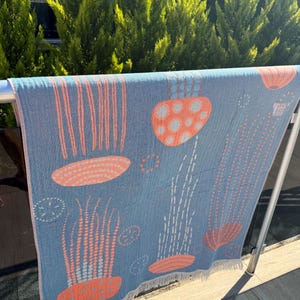 K&ouml;nnte beinhalten: Ein blaues Strandtuch mit einem Quallen-Design in Koralle und Wei&szlig;. Das Tuch h&auml;ngt drau&szlig;en an einer Metallstange. Die Quallen haben gepunktete Tentakel und glockenf&ouml;rmige K&ouml;rper. Das Tuch hat einen Fransenrand.