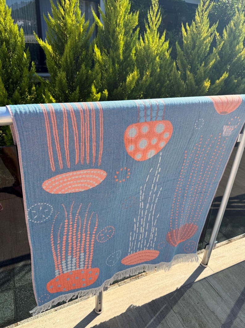 K&ouml;nnte beinhalten: Ein blaues Strandtuch mit einem Quallen-Design in Koralle und Wei&szlig;. Das Tuch h&auml;ngt drau&szlig;en an einer Metallstange. Die Quallen haben gepunktete Tentakel und glockenf&ouml;rmige K&ouml;rper. Das Tuch hat einen Fransenrand.