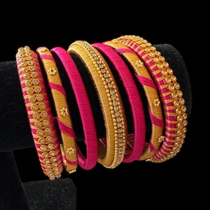 May include: A collection of gold and pink bangles. Some bangles are solid pink, while others have gold accents and embedded gemstones. The bangles are displayed on a black stand against a black background.