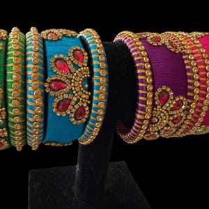 May include: A collection of colorful bangles in shades of green, blue, purple, and pink. Each bangle is adorned with intricate gold-colored embellishments and sparkling red and green gemstones. The bangles are displayed on a black stand against a black background.
