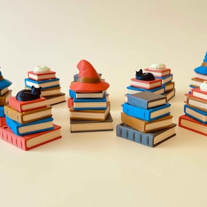 May include: A collection of miniature stacks of books with various toppers. Some stacks have a black cat figurine, a witch hat, or a small cake. The books are in shades of blue, brown, and red, with white pages.