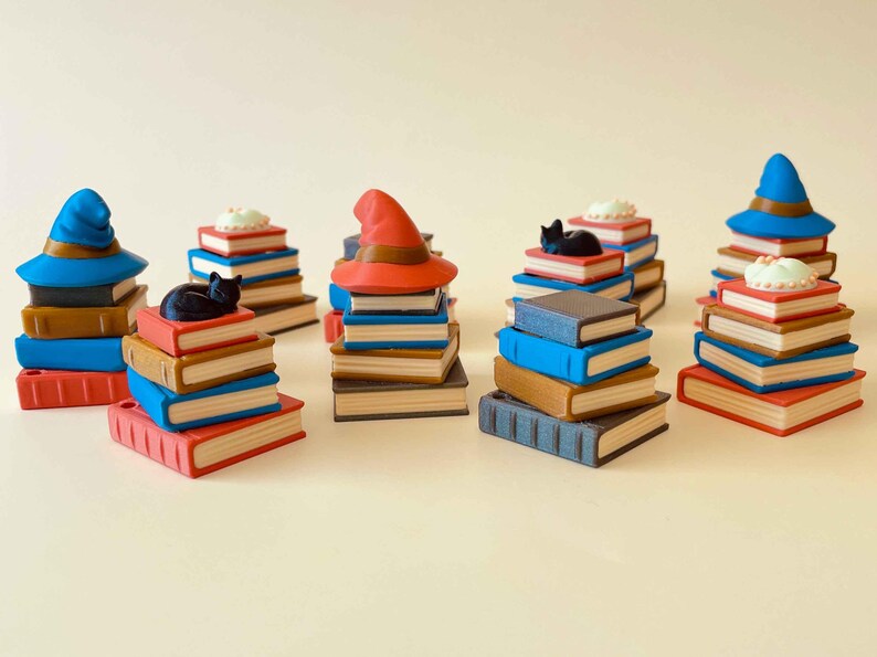 May include: A collection of miniature stacks of books with various toppers. Some stacks have a black cat figurine, a witch hat, or a small cake. The books are in shades of blue, brown, and red, with white pages.