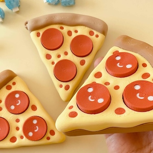 Puede incluir: Tres porciones de pizza con corteza amarilla, bordes de corteza marr&oacute;n y aderezos de pepperoni rojos. El pepperoni tiene detalles blancos. Las porciones de pizza est&aacute;n sobre una superficie amarillo claro. Peque&ntilde;os objetos azules y blancos en el fondo.