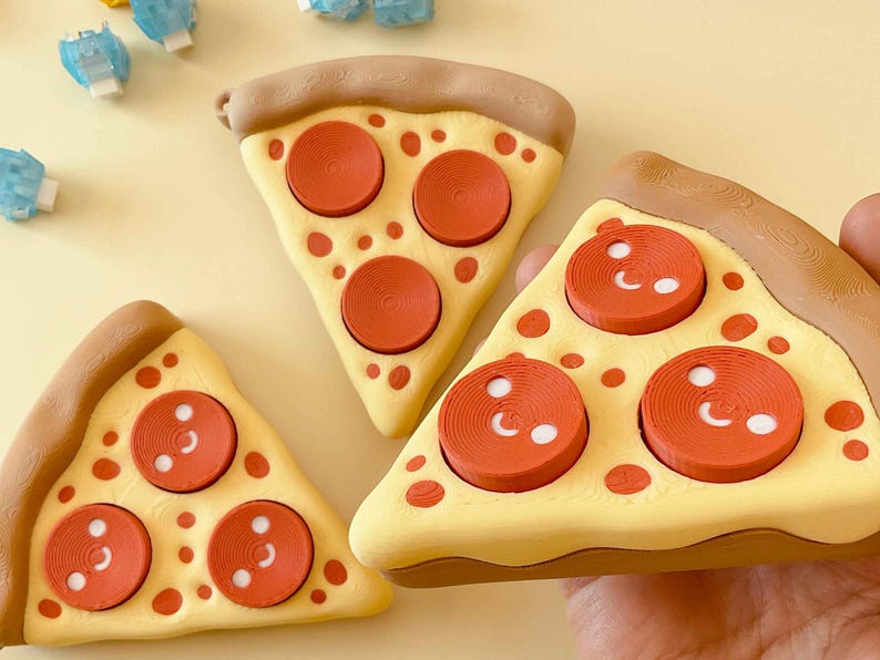 Puede incluir: Tres porciones de pizza con corteza amarilla, bordes de corteza marr&oacute;n y aderezos de pepperoni rojos. El pepperoni tiene detalles blancos. Las porciones de pizza est&aacute;n sobre una superficie amarillo claro. Peque&ntilde;os objetos azules y blancos en el fondo.