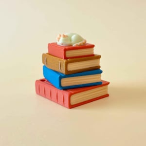 May include: A stack of four miniature books in red, brown, blue, and red, with a small white cat figurine sleeping on top. The books have cream-colored pages and are stacked on a light beige surface.