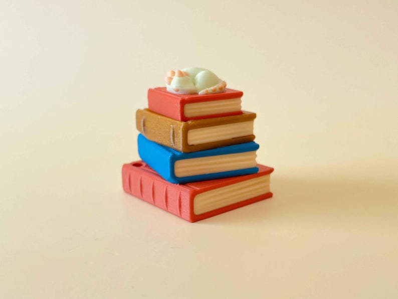 May include: A stack of four miniature books in red, brown, blue, and red, with a small white cat figurine sleeping on top. The books have cream-colored pages and are stacked on a light beige surface.