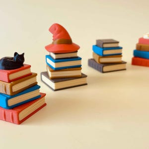 May include: A collection of miniature book stacks in various colors, including red, blue, and brown. One stack features a black cat figurine, while another has a red witch hat. The theme suggests a fantasy or literary concept.