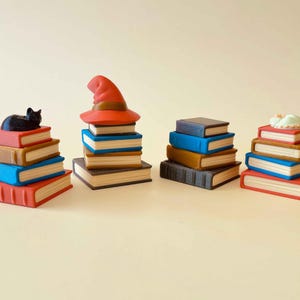 May include: Several miniature book stacks in shades of red, blue, and brown. Some are adorned with a black cat, a witch's hat, or a sleeping figure. The books have white page edges.