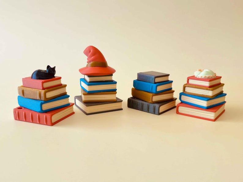 May include: A collection of miniature book stacks in various colors, including red, blue, and brown. Some stacks are topped with a black cat, a witch's hat, or a sleeping figure. The books have white page edges.