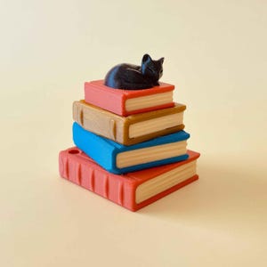 May include: A stack of colorful books with a black cat figurine resting on top. The books are red, brown, and blue, with white pages. The cat is curled up, adding a whimsical touch to this decorative item.