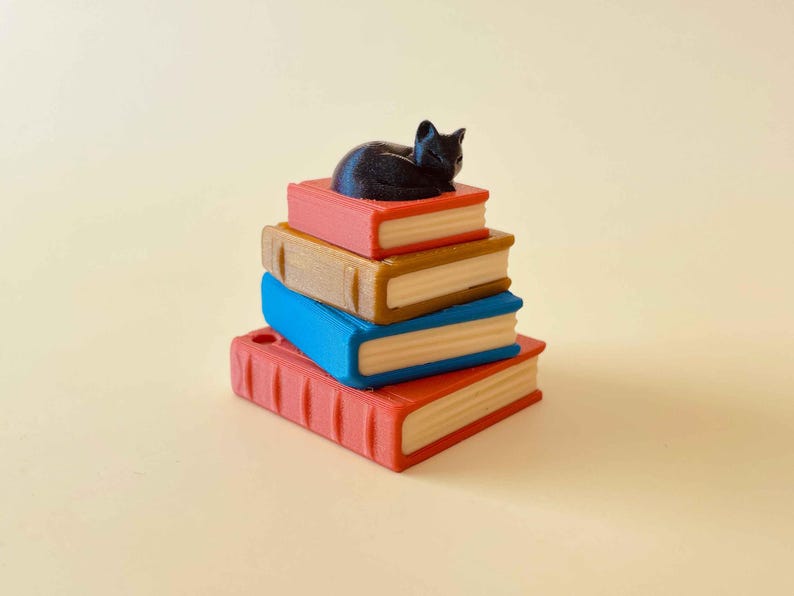 May include: A stack of colorful books with a black cat figurine resting on top. The books are red, brown, and blue, with white pages. The cat is curled up, adding a whimsical touch to this decorative item.