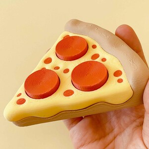 Puede incluir: Una porci&oacute;n de pizza con base amarilla, pepperoni rojo y corteza marr&oacute;n. La porci&oacute;n de pizza se sostiene en una mano. El pepperoni es circular y elevado. La corteza tiene un aspecto texturizado.