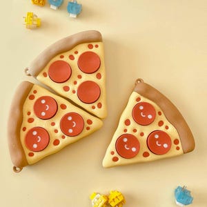 Puede incluir: Tres accesorios con forma de porci&oacute;n de pizza con pepperoni rojo y un dise&ntilde;o de cara sonriente. La corteza es marr&oacute;n y la base es amarilla. Peque&ntilde;os accesorios coloridos est&aacute;n esparcidos alrededor de las porciones de pizza sobre un fondo amarillo.