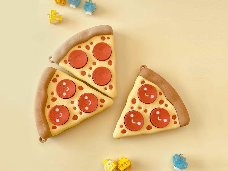 Puede incluir: Tres accesorios con forma de porci&oacute;n de pizza con pepperoni rojo y un dise&ntilde;o de cara sonriente. La corteza es marr&oacute;n y la base es amarilla. Peque&ntilde;os accesorios coloridos est&aacute;n esparcidos alrededor de las porciones de pizza sobre un fondo amarillo.
