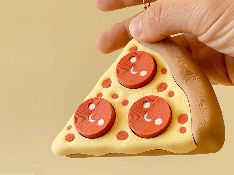 Puede incluir: Un llavero con forma de porci&oacute;n de pizza con tres rodajas de pepperoni rojas y detalles blancos. La corteza es marr&oacute;n y el queso es amarillo. El fondo es de color amarillo claro.