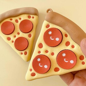 Puede incluir: Dos adornos de rebanadas de pizza. Cada rebanada tiene una base amarilla con pepperoni rojo y una corteza marr&oacute;n. Los pepperoni tienen detalles blancos. Una rebanada es sostenida por una mano. Los adornos son para un &aacute;rbol de Navidad.