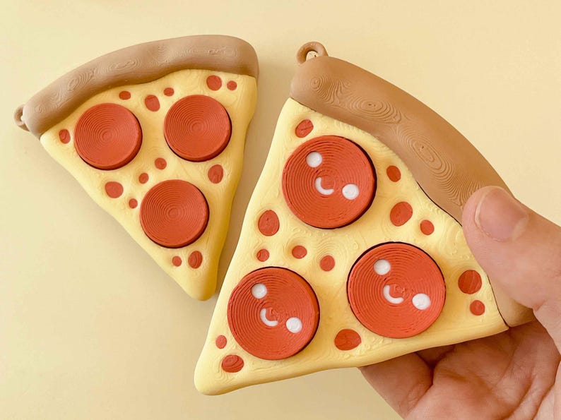 Puede incluir: Dos adornos de rebanadas de pizza. Cada rebanada tiene una base amarilla con pepperoni rojo y una corteza marr&oacute;n. Los pepperoni tienen detalles blancos. Una rebanada es sostenida por una mano. Los adornos son para un &aacute;rbol de Navidad.