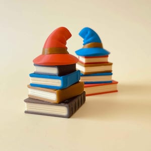 May include: Two stacks of books topped with wizard hats. One stack has a red hat, the other a blue hat. The books are various colors, including brown, blue, and black, with white pages. The hats have brown bands.