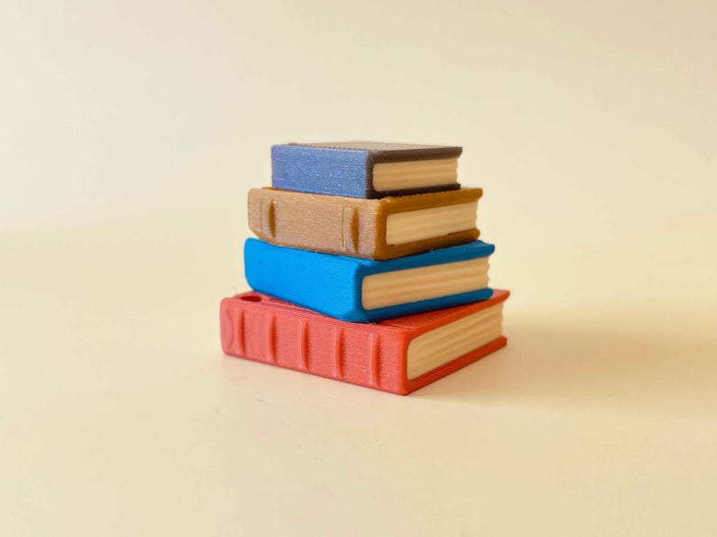 May include: A stack of four miniature books in red, blue, tan, and dark blue. The books are stacked on top of each other, with the red book at the bottom and the dark blue book at the top. The pages are a light tan color.