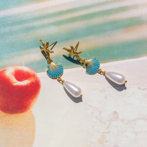 Peut inclure: Boucles d'oreilles dorées avec une coquille de mer bleue et un charme d'étoile de mer, suspendues à une goutte de perle blanche.