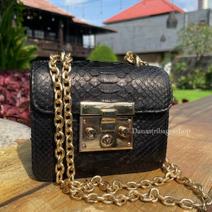 Pode incluir: Bolsa preta com corrente dourada e fecho dourado. A bolsa tem um padrão texturizado semelhante à pele de cobra. A bolsa está sobre uma superfície de madeira.
