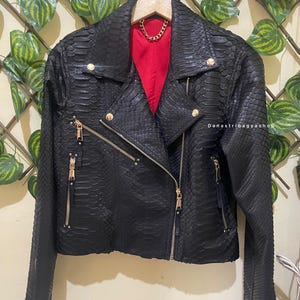 Puede incluir: Chaqueta negra de cuero sintético con textura de piel de serpiente, cremalleras y botones dorados. La chaqueta tiene un forro rojo y un detalle de cadena dorada. La chaqueta está colgada en una percha de madera.