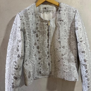 Puede incluir: Chaqueta blanca y gris con estampado de piel de serpiente y cremallera dorada. La chaqueta tiene cuello redondo y mangas largas. El forro es blanco. La chaqueta está colgada en una percha de madera.